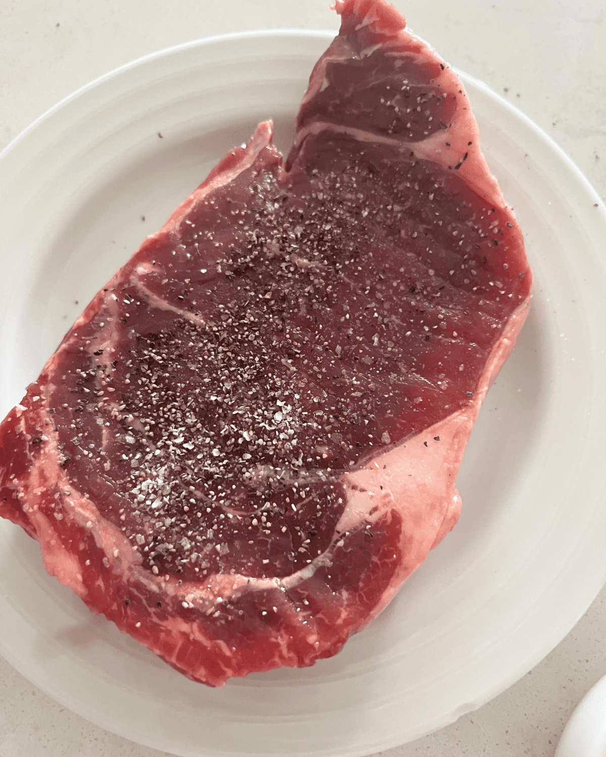 seasoned steak with salt and pepper on a white plate.