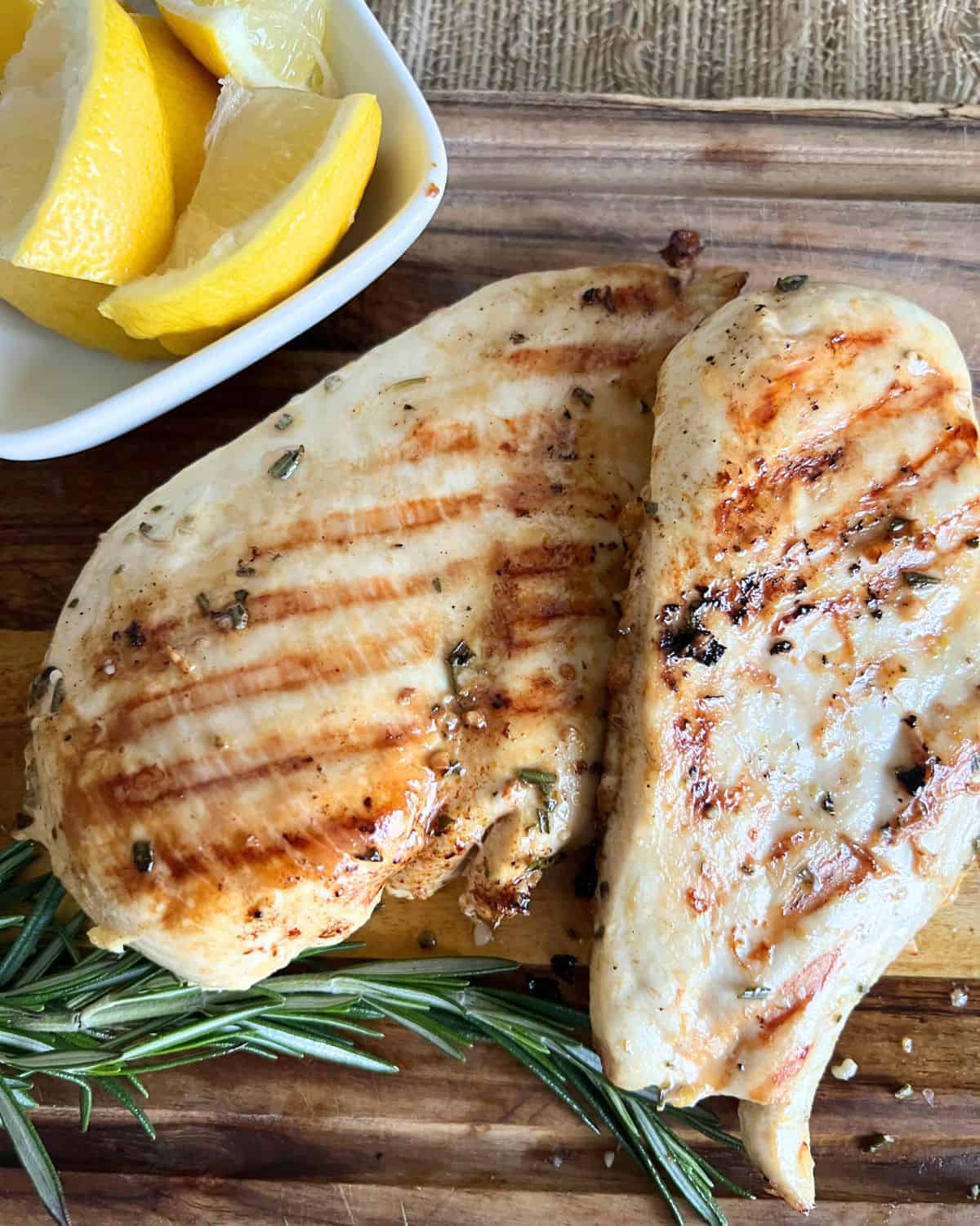 Finished grilled lemon chicken breasts on a cutting board next to lemon wedges and rosemary. 