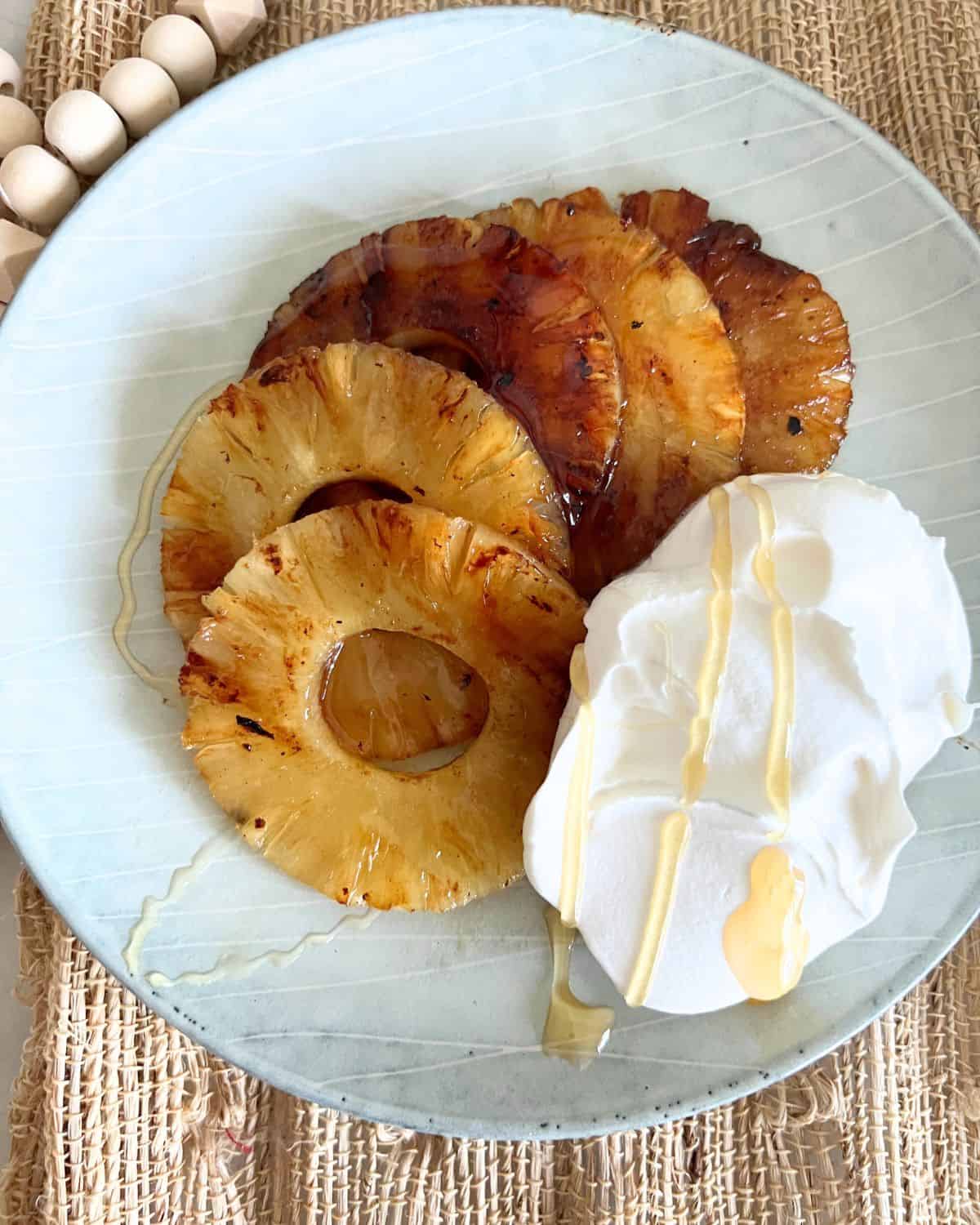 Sliced grilled pineapple next to cool whip on a blue plate drizzled in honey. 