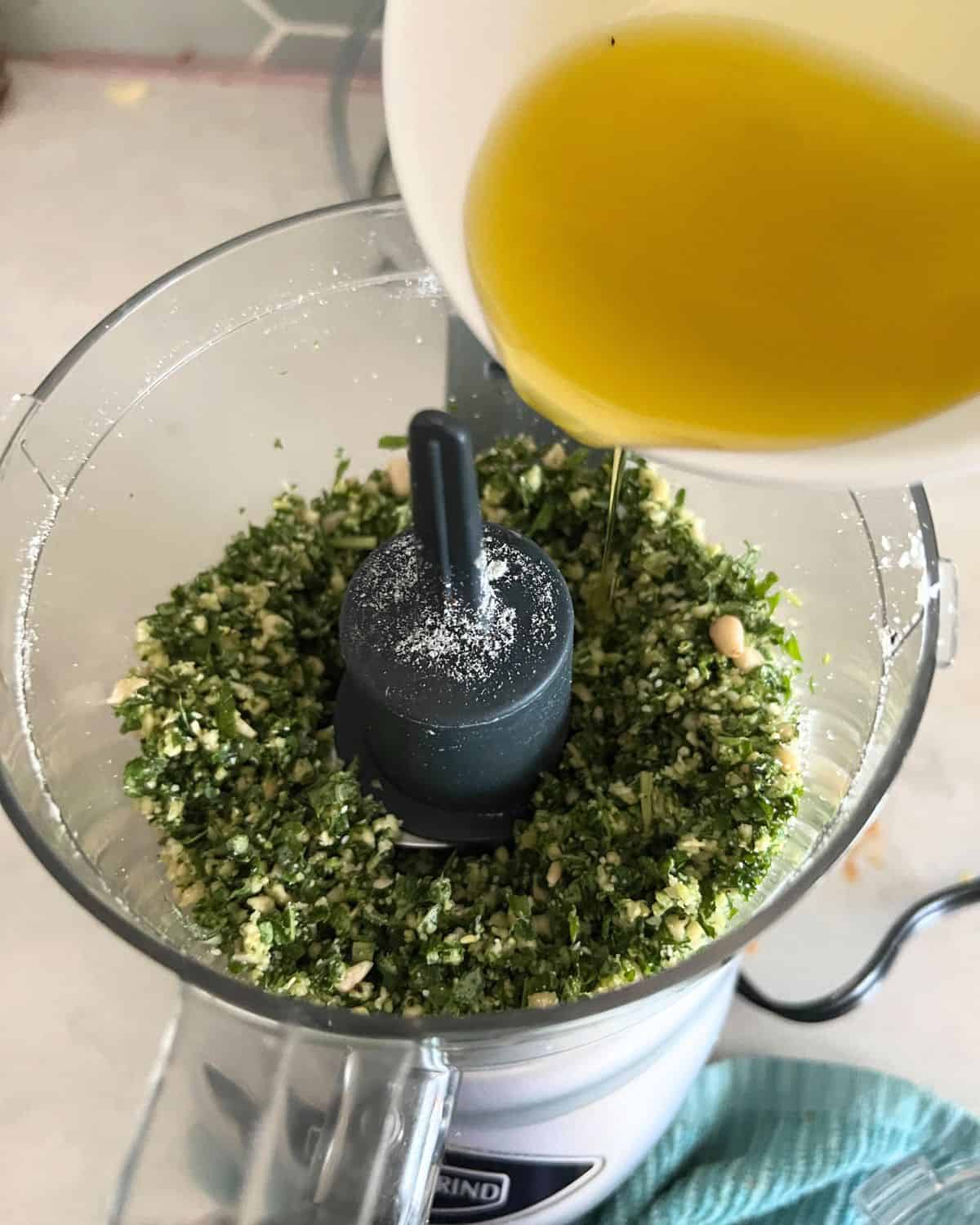 Adding olive oil to pesto ingredients in a food processor. 