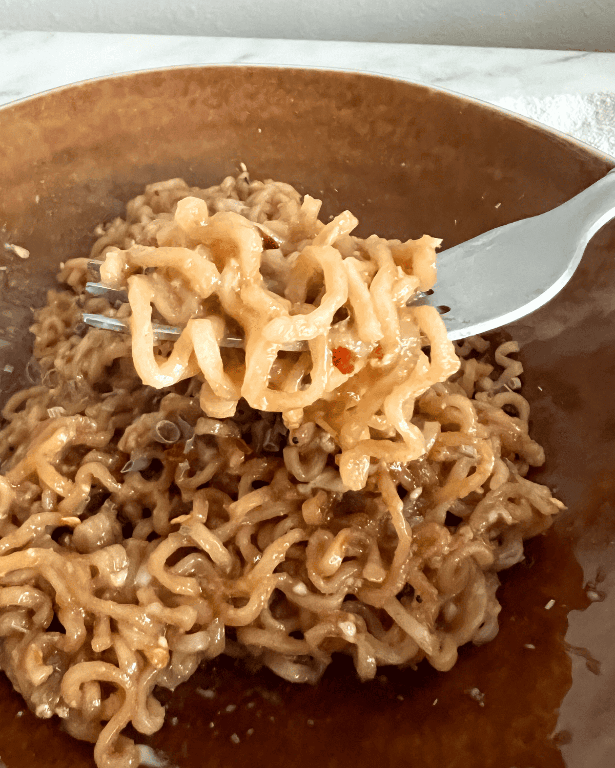 a fork holding ramen noodle over a brown dish of noodles. 