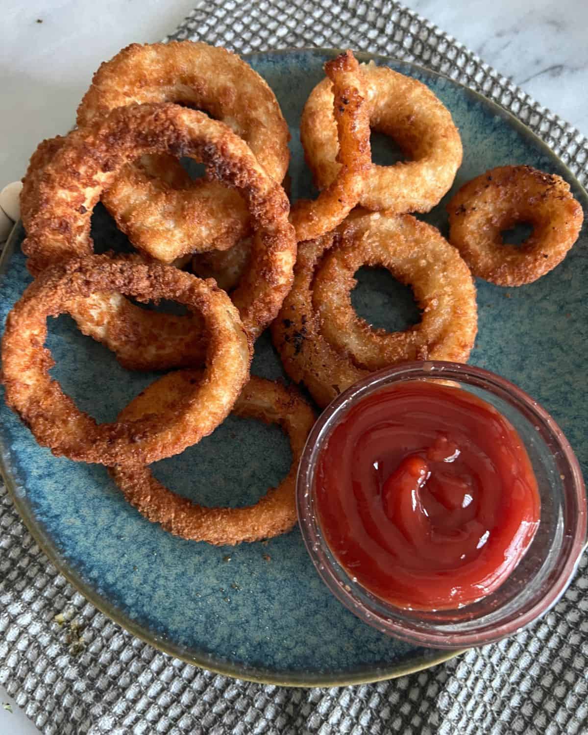 Crispy air fried onion rings on a blue plate next to ketchup. 