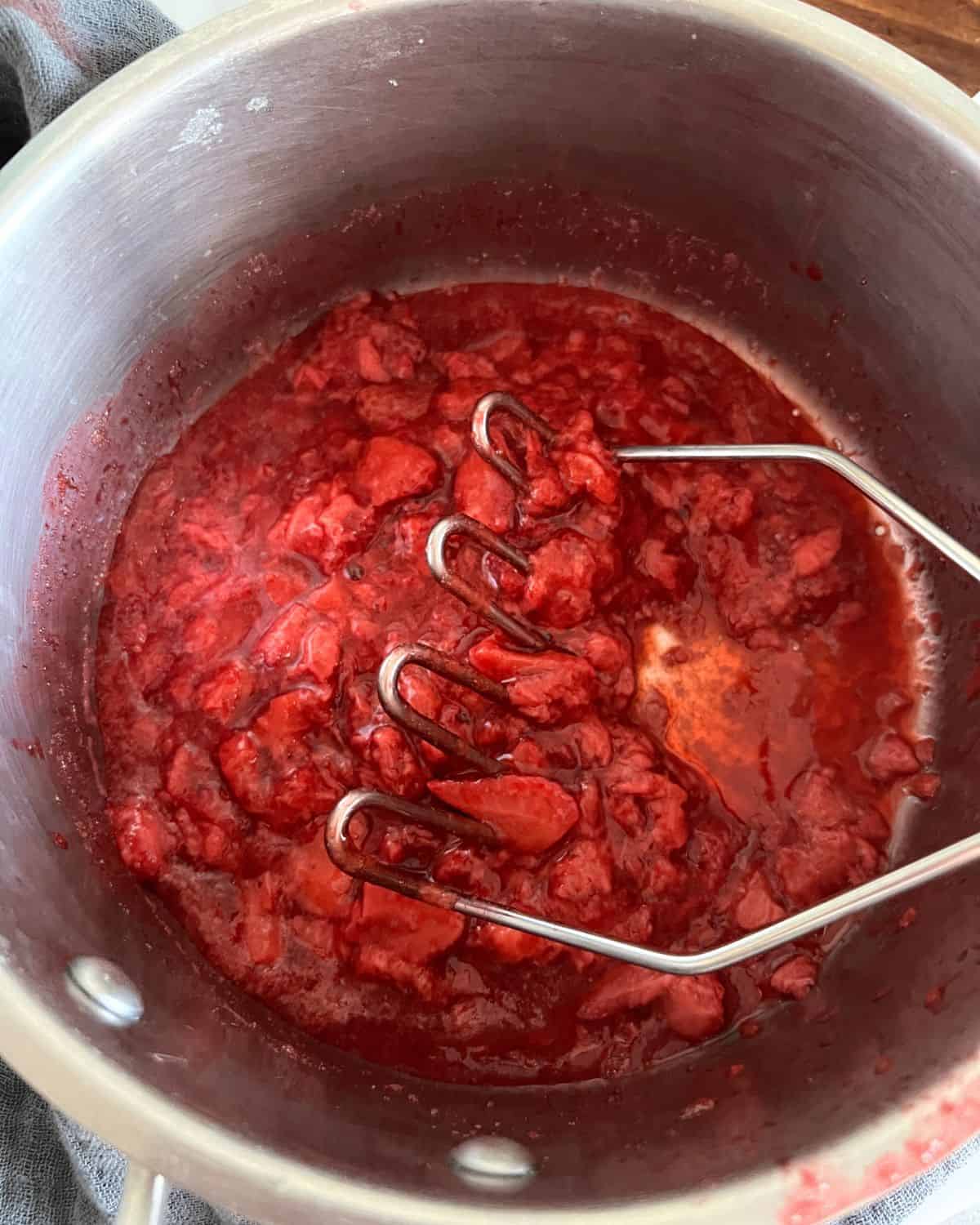 Mashed strawberries in a pan with a potato masher.