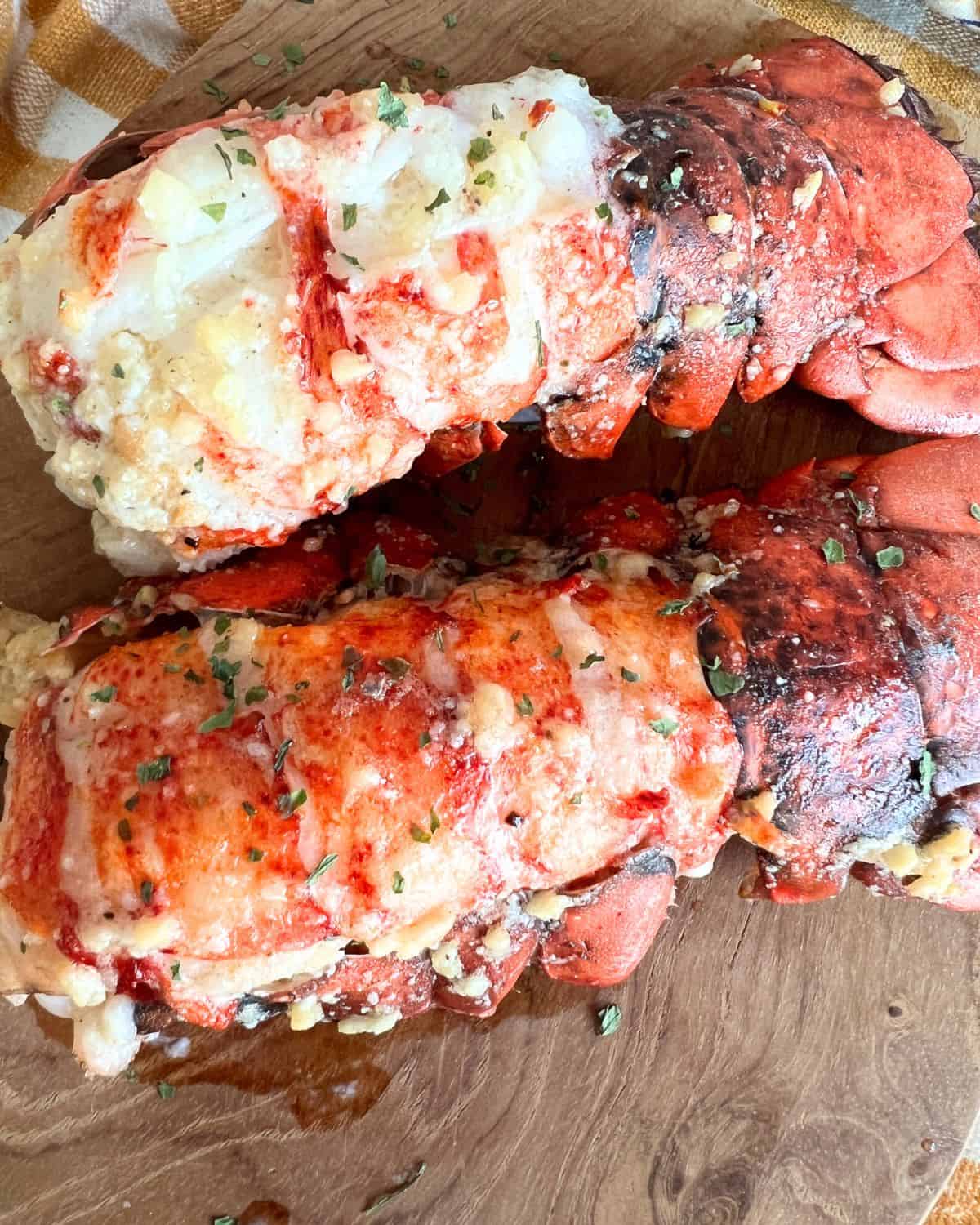 Baked lobster tails coated in garlic butter sauce on a wooden cutting board. 