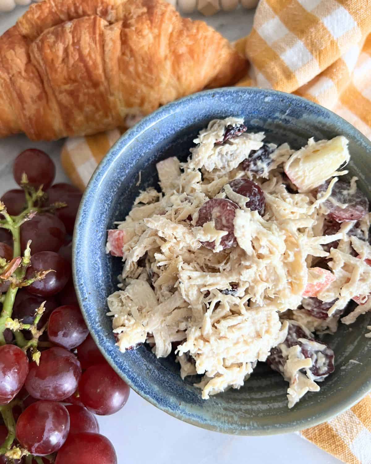 Chicken salad with grapes in a blue bowl next to grapes and a croissant.