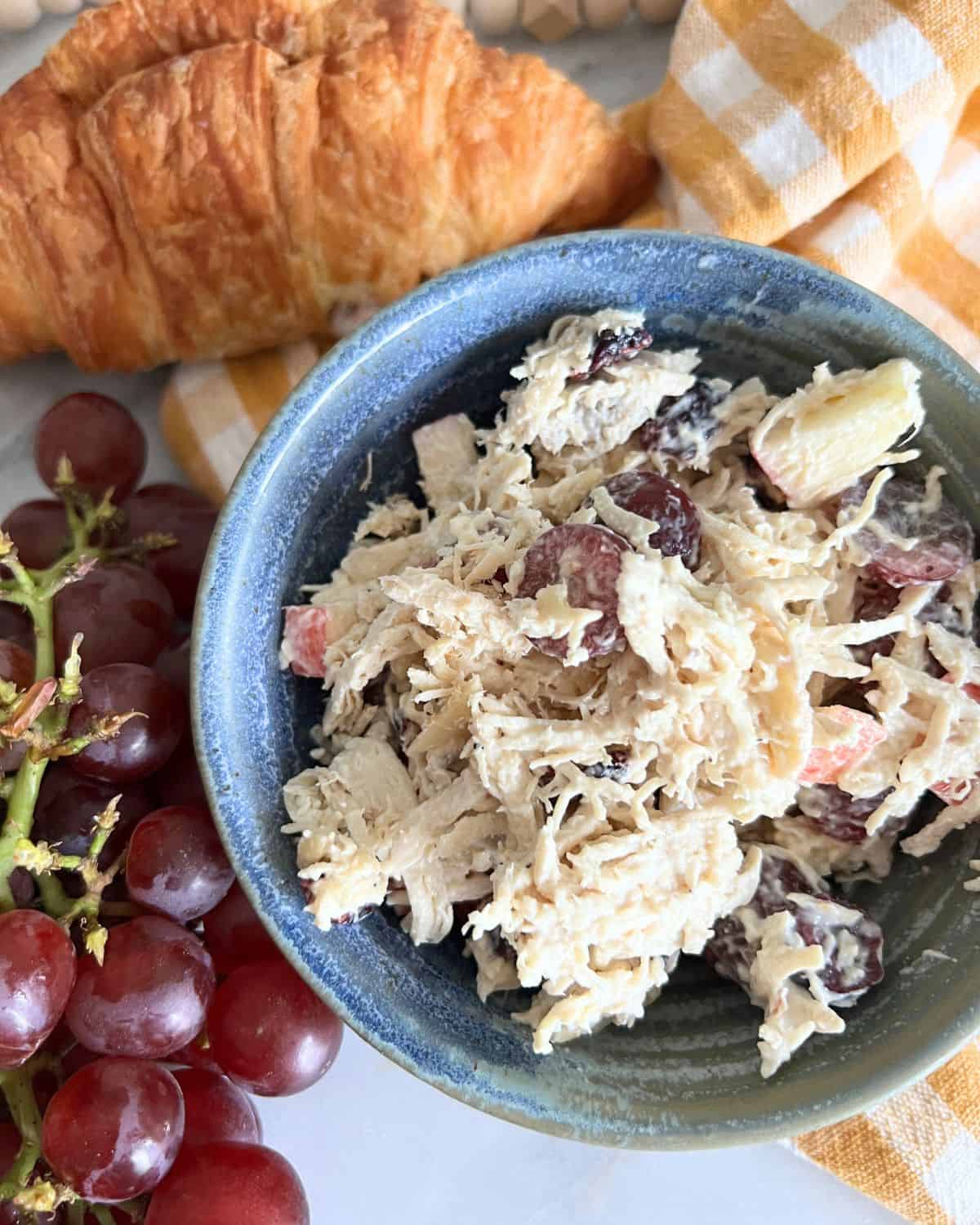 Chicken salad with grapes in a blue bowl next to grapes and a croissant. 