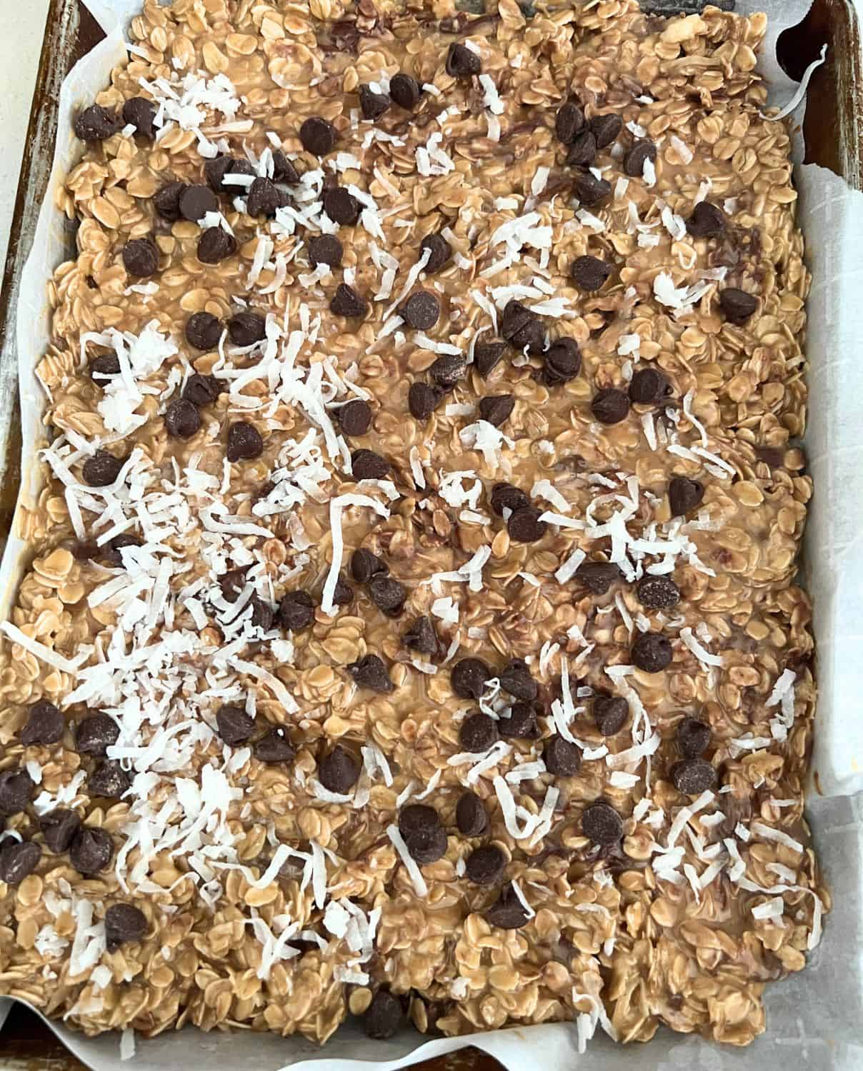 Granola mixture pressed down on a pan and topped with chocolate chips and coconut. 