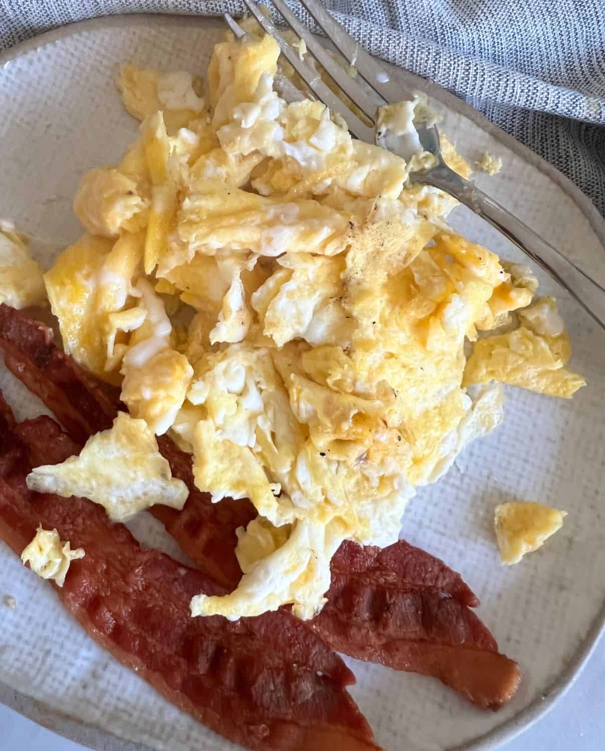 Cooked scrambled eggs next to some bacon on a white plate. 