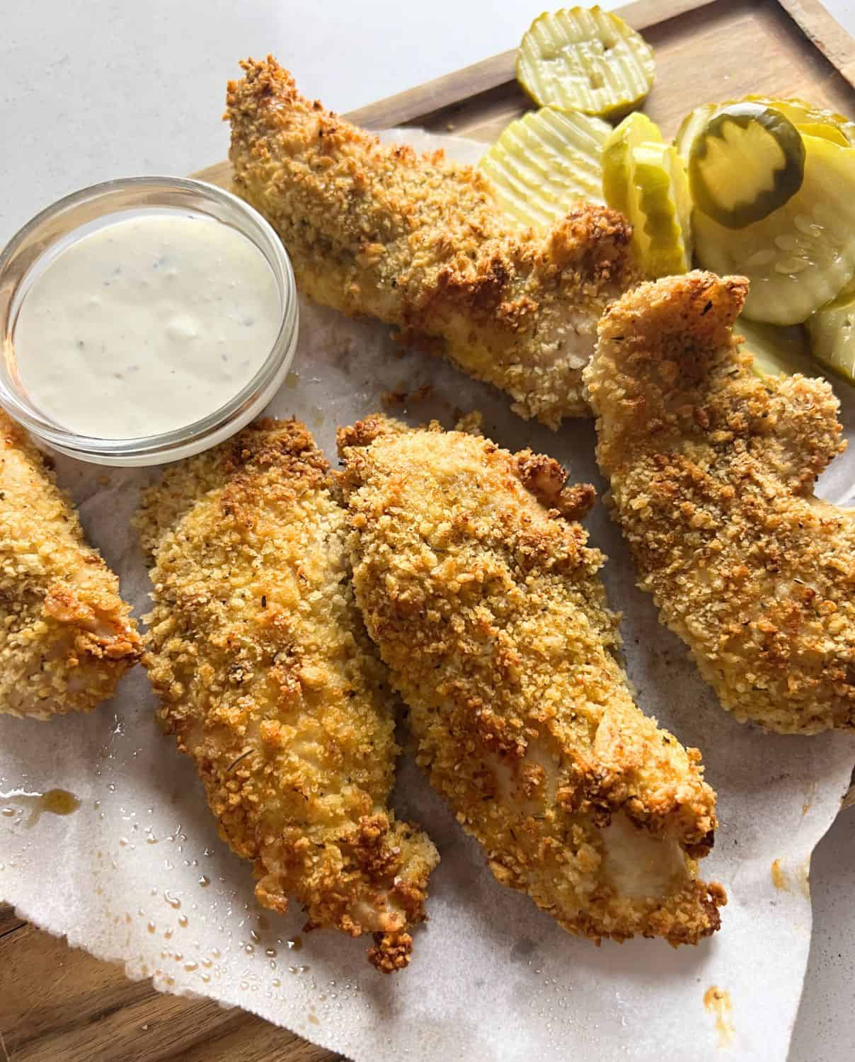 Air fried chicken tenders next to ranch and pickles on a cutting board. 