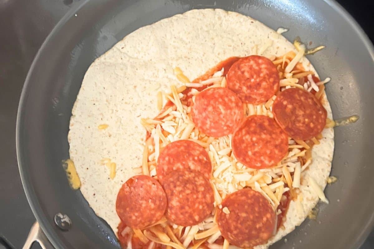 Tortilla in a skillet topped with pizza sauce, cheese, and pepperoni.