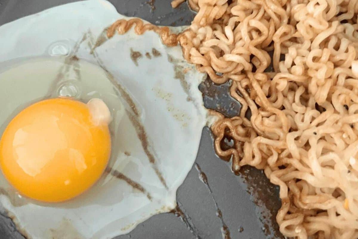 Cracked egg in a skillet next to ramen noodles.
