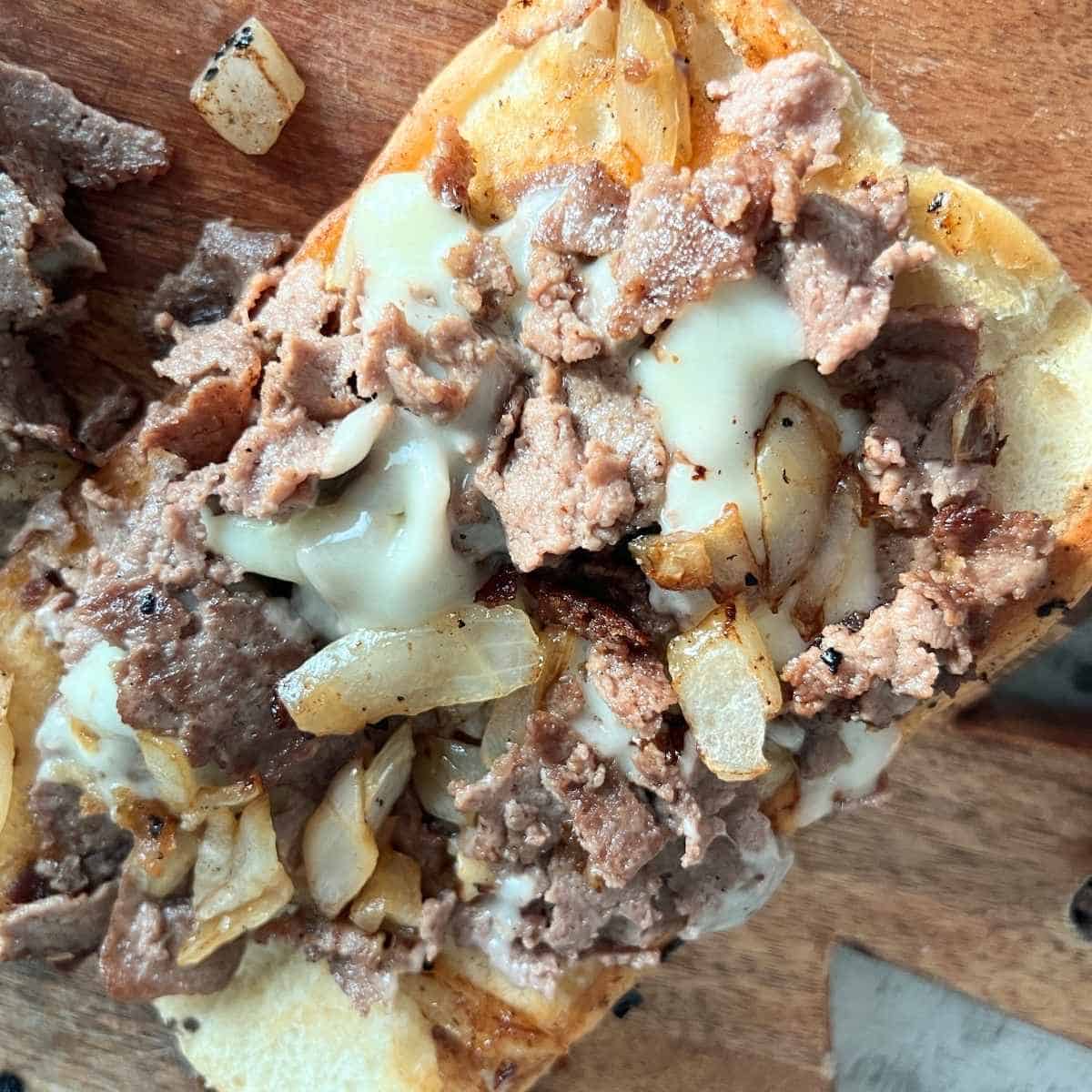 Close-up of a Philly cheesesteak sandwich with thin-sliced ribeye steak, melted provolone cheese, and sautéed onions stuffed inside a toasted hoagie roll, served on a wooden board.