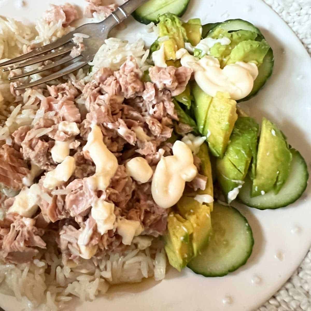 A colorful tuna rice bowl topped with sliced avocado and fresh cucumber on a bed of white rice, drizzled with sesame oil and soy sauce, and garnished with sesame seeds and green onion in a white ceramic bowl.